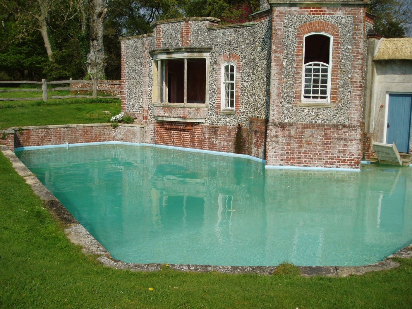 Turquoise walled pool reflecting an old red brick and flint building with arched windows, water sits calm against the facade, surrounded by trimmed grass, a wooden fence and trees.