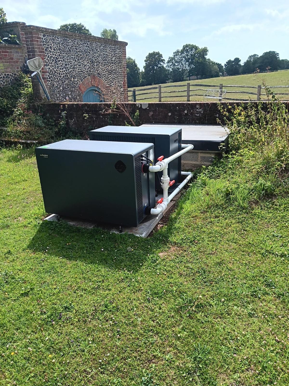 Two dark equipment units connected by white PVC pipes sit on a concrete pad in a sunny grassy yard beside a flint-and-brick wall with a fenced pasture and trees beyond.