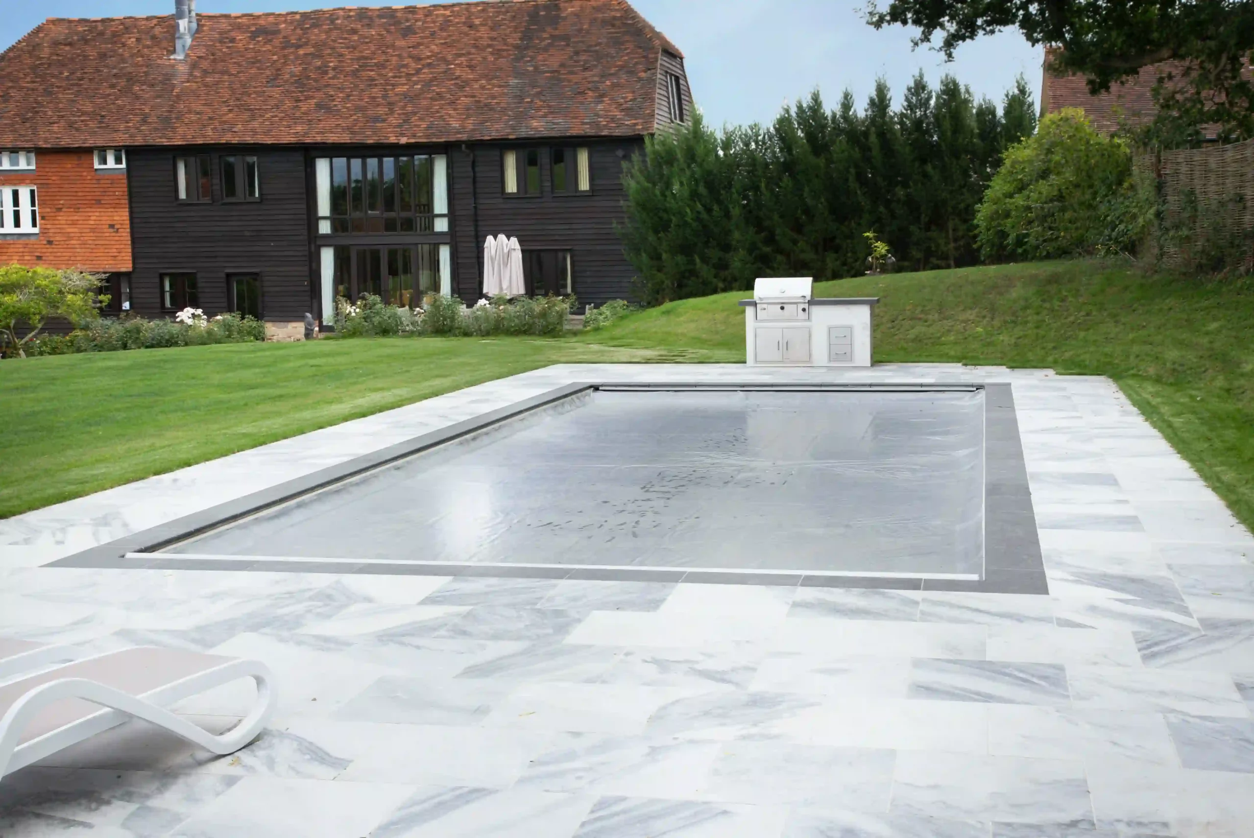 Rectangular in-ground swimming pool sits covered by a taut gray safety cover, surrounded by white marble tiles, with a manicured lawn, stainless grill and dark wooden farmhouse in the background.